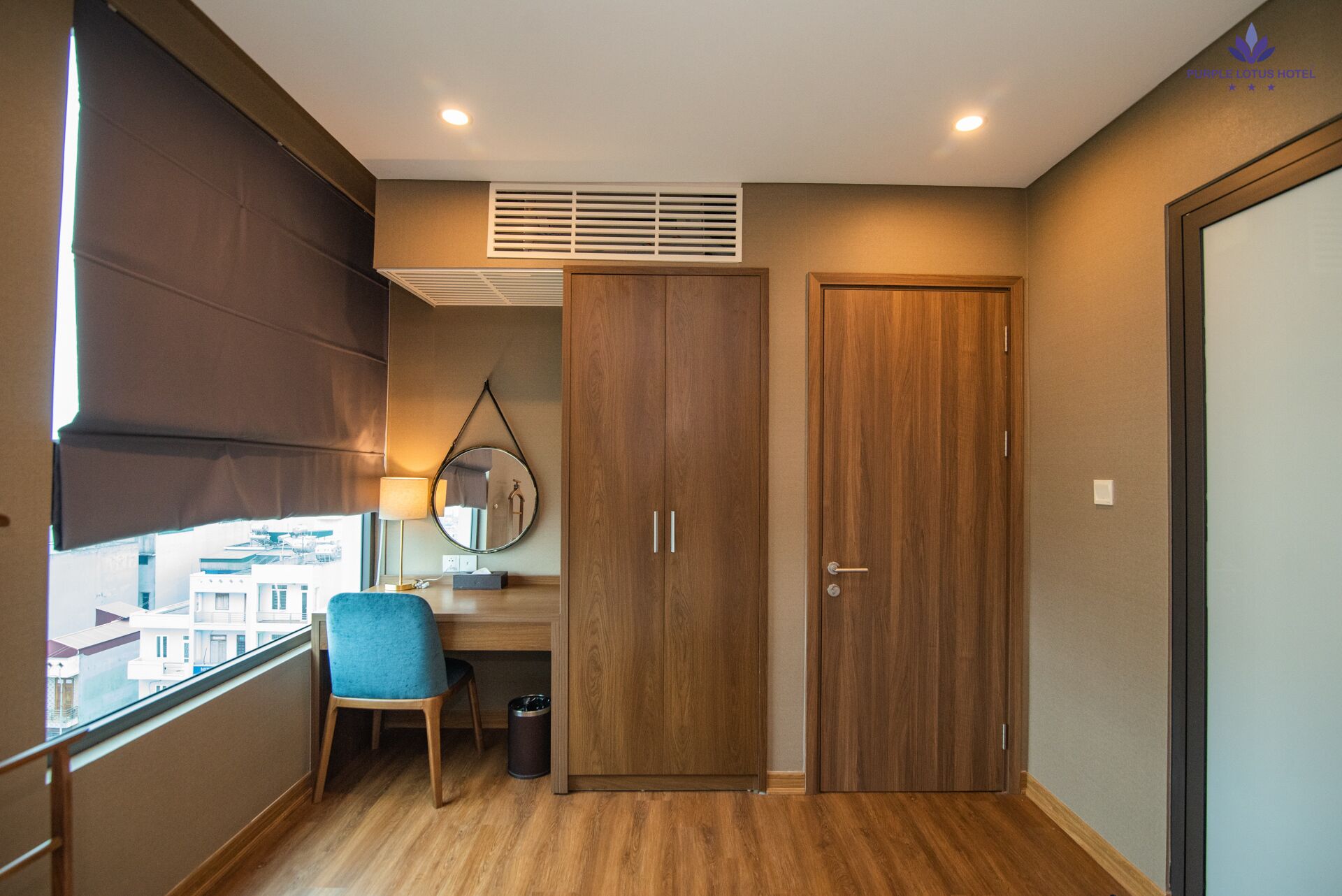 A hotel room with a wooden floor, a desk with a chair, a mirror, and a wardrobe.