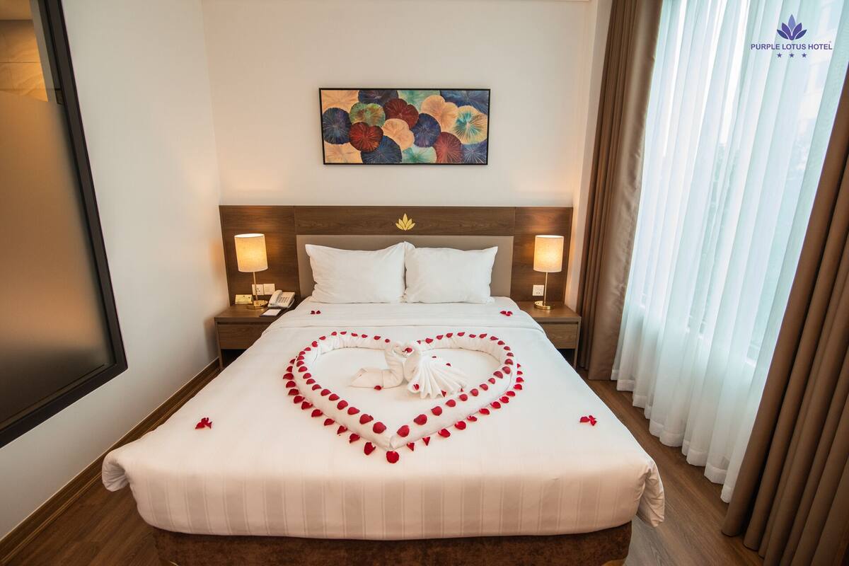Une chambre d’hôtel avec un lit, deux tables de chevet, un tableau et une fenêtre avec des rideaux.
