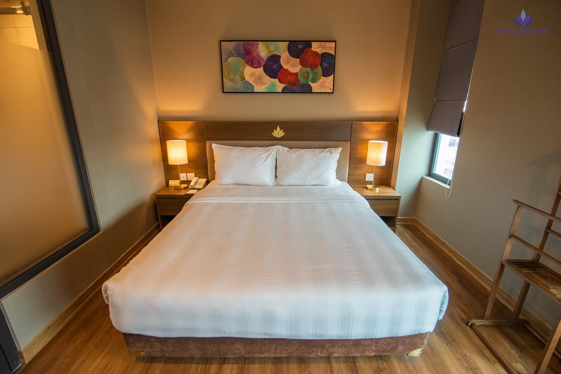 A large, neatly made bed with white linens in a hotel room. There are two bedside tables with lamps, a wall-mounted painting above the bed, and a window on the right side.