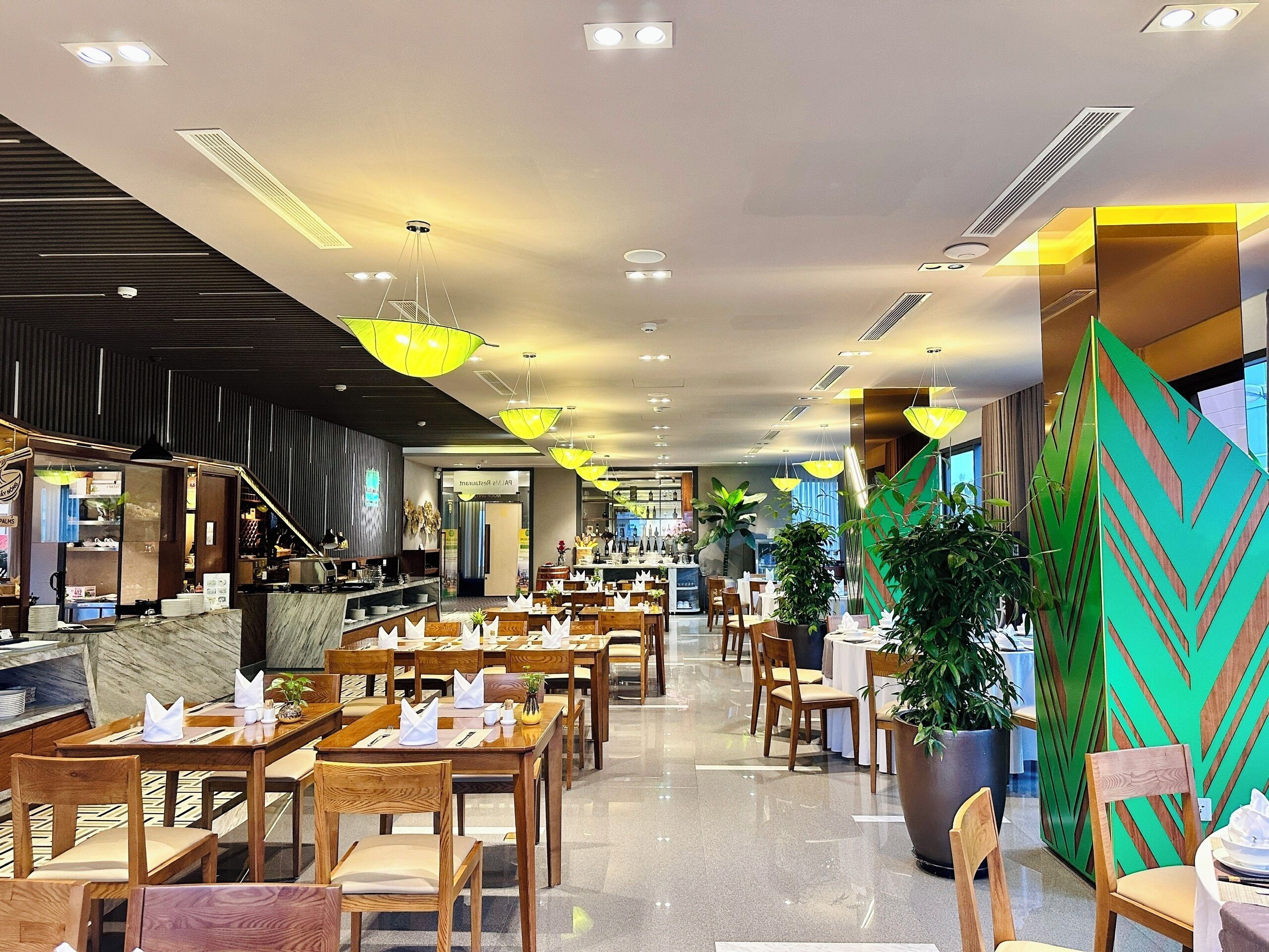 A modern restaurant with wooden tables and chairs, bright pendant lights, and potted plants.