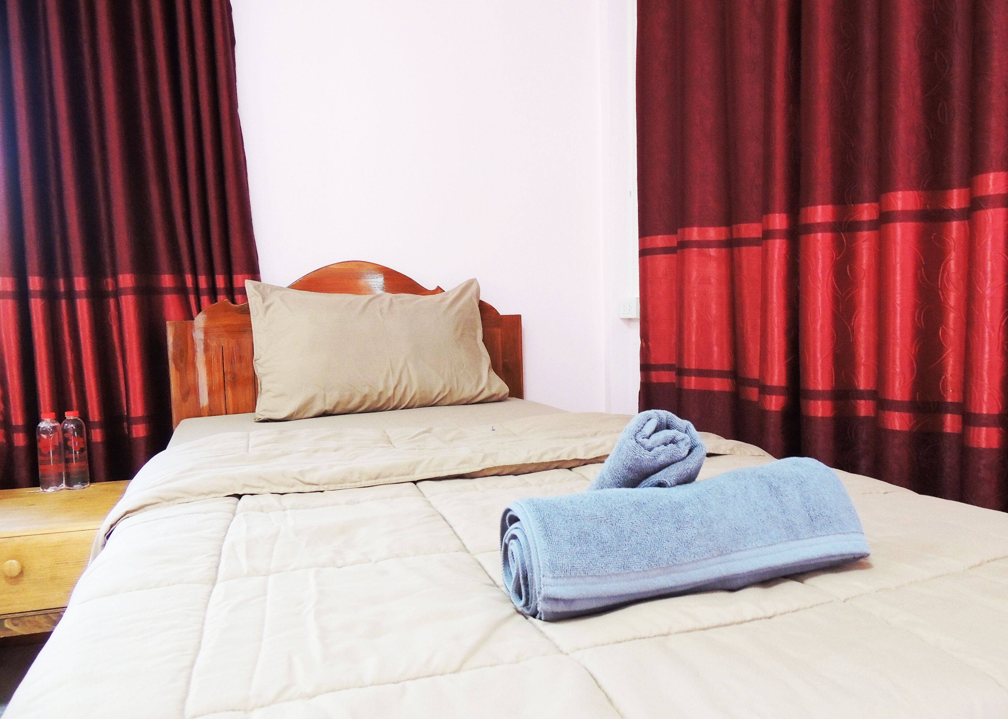 A neatly made bed with a beige comforter and pillows, a wooden headboard, and red curtains.