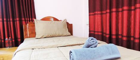 A neatly made bed with a beige comforter and pillows, a wooden headboard, and red curtains.