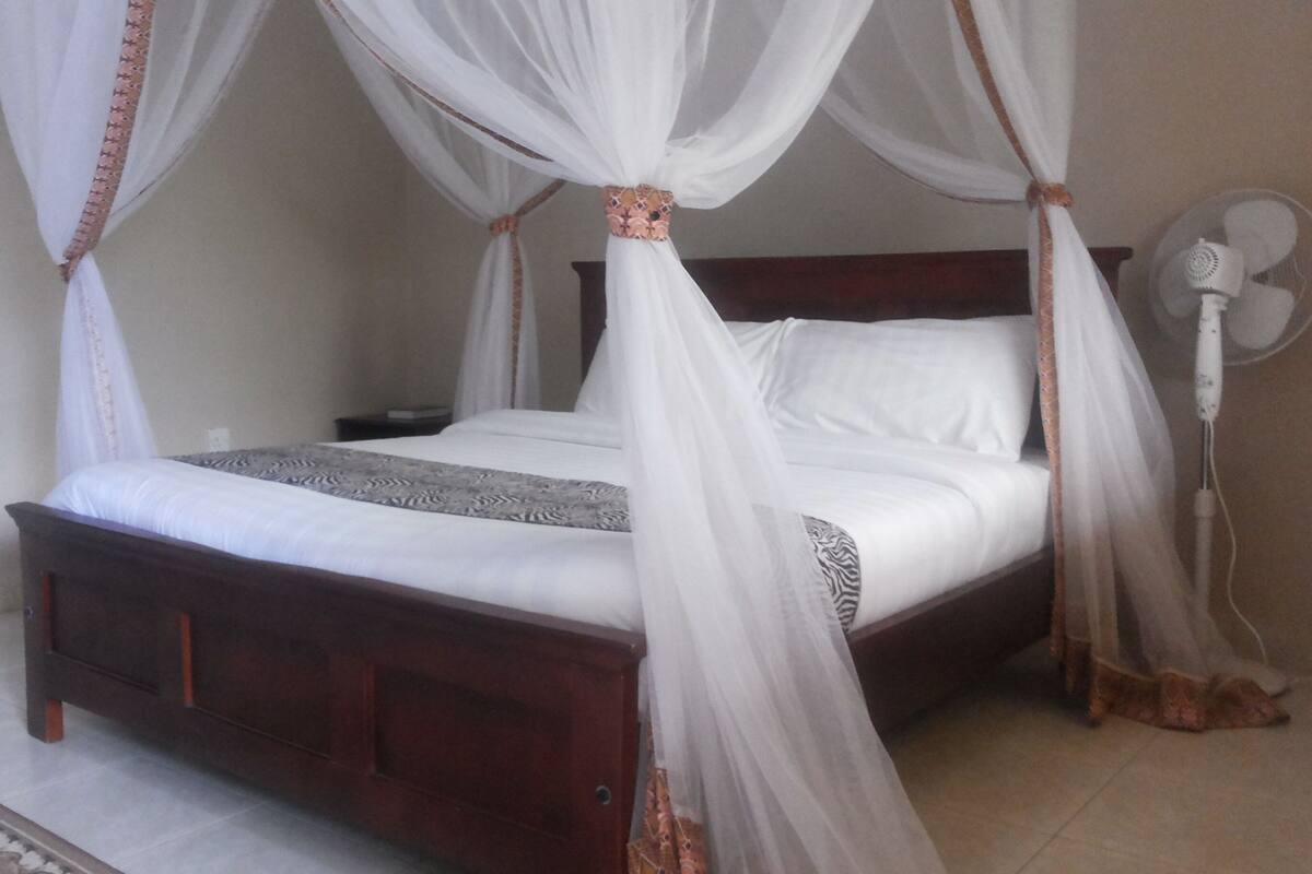 A bed with a wooden frame and white bedding, a bedside table, a wall-mounted fan, and a patterned rug on the floor.