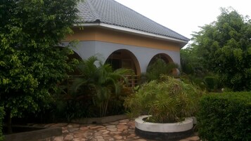 Un edificio con techo de tejas, ventanas en arco y un sendero de piedra que conduce a una jardinera circular con plantas tropicales.