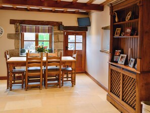 A dining area with a wooden table and chairs, a stone wall, a television mounted on the wall, and a wooden shelf with framed pictures. - 3 bedroom accommodation in Hebden, near Grassington (Hebden, near Grassington)