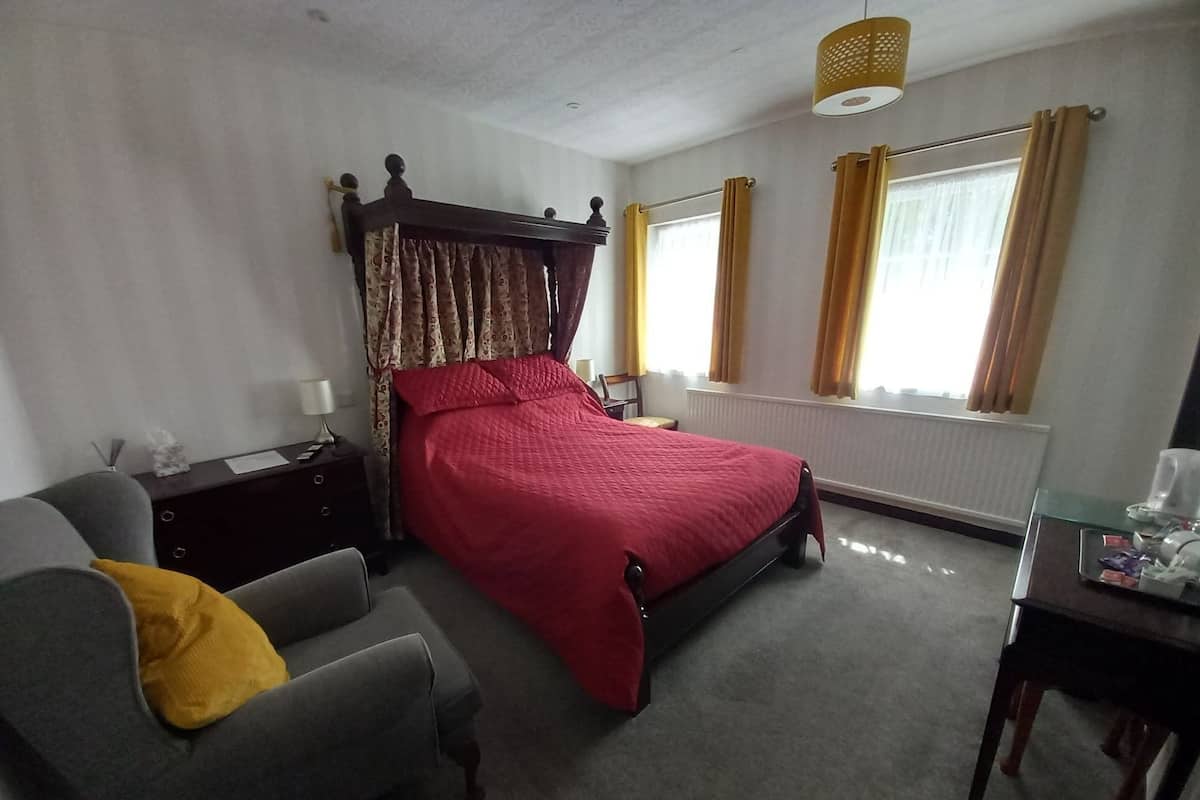 Quarto com cama de dossel, poltrona cinza com manta amarela, mesa de cabeceira com abajur e janela com cortinas.