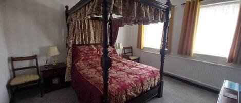 Una cama con dosel adornada con una colcha estampada, una mesita de noche de madera, una silla y una ventana con cortinas.