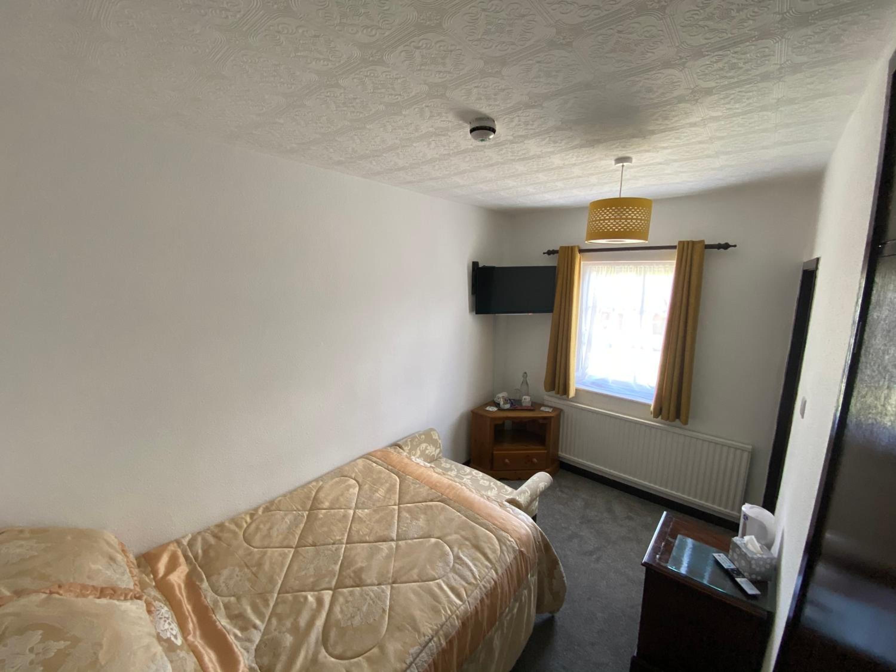 Una camera d'albergo con un letto, un comodino, una finestra con tende, una televisione a muro e una telecamera di sicurezza a soffitto.