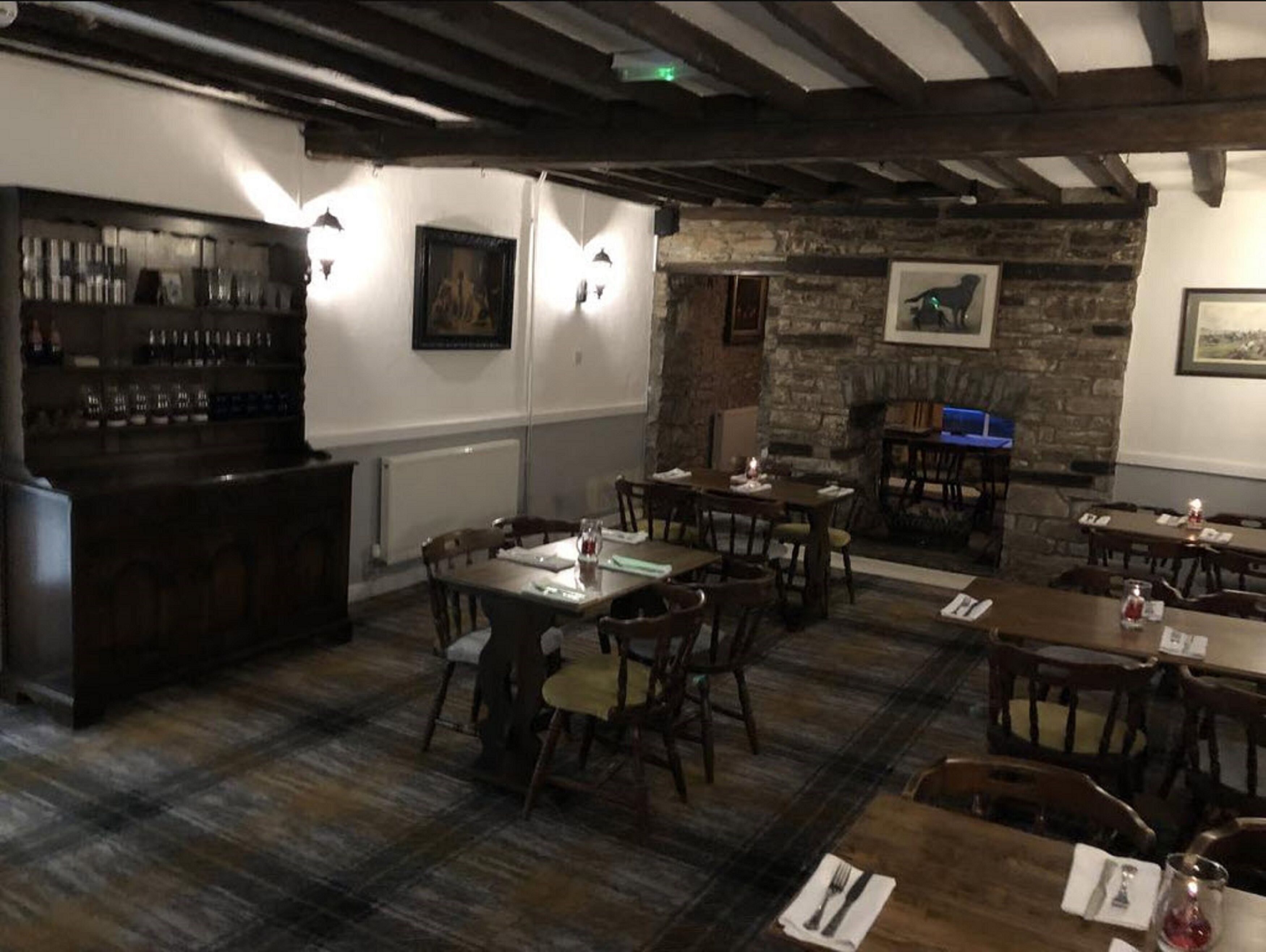 Un pub rústico con vigas de madera a la vista, una pared de piedra y una zona de bar con botellas. Mesas preparadas para comer.
