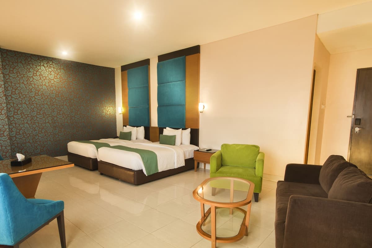 Une chambre d’hôtel avec un lit, deux tables de chevet, un fauteuil vert, un canapé marron, une table basse en verre et un mur à motifs.