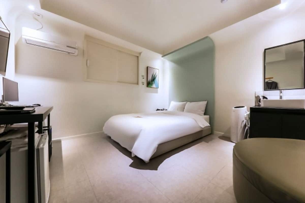 A modern hotel room with a bed, a desk with a computer, a bedside table, a mirror, and a bathroom with a sink and toilet.