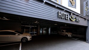 Ein Hotel Eingang mit modernem Design, mit einer großen Schilder, auf der 'HOTEL YAM' steht. Vorne sind zwei Autos geparkt, und der Eingang ist von einem Vordach überdacht.
