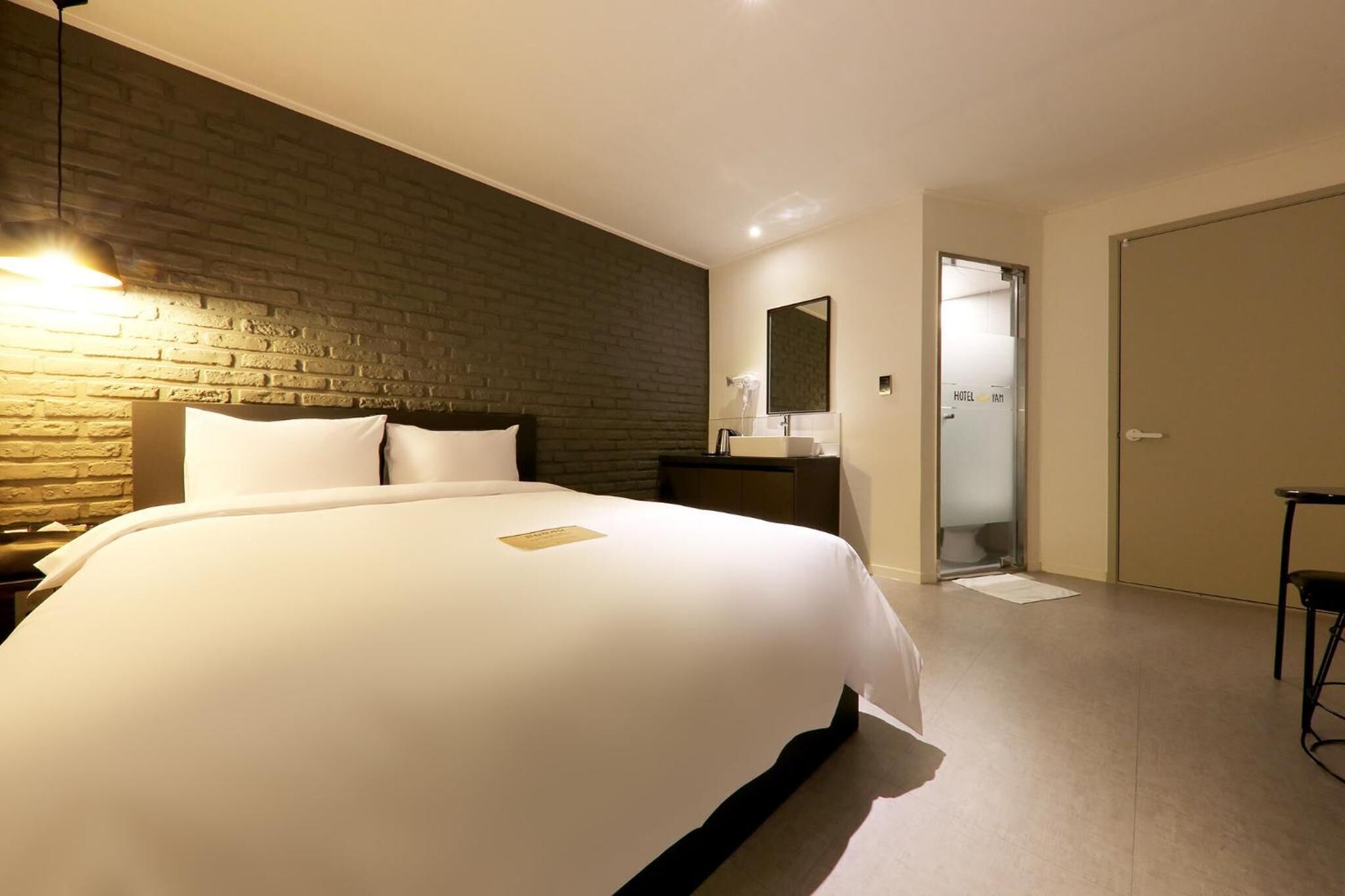 A modern hotel room with a large bed, a brick accent wall, a bedside table, a mirror, and a door leading to a bathroom.