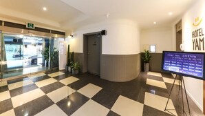Eine Hotelhalle mit Schachbrettmuster-Boden, einer Rezeption, einer digitalen Anzeige und einem Glasgang zum Parkplatz.