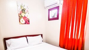 A bedroom with a wooden bed, white bedding, a floral artwork on the wall, an air conditioning unit, and a window with red curtains. - Downtown Suites (Cagayan de Oro)