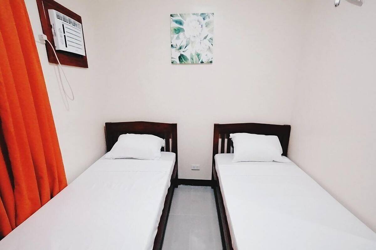 Quarto com duas camas de solteiro, uma janela pequena com vista para flores e um ar condicionado fixado na parede.