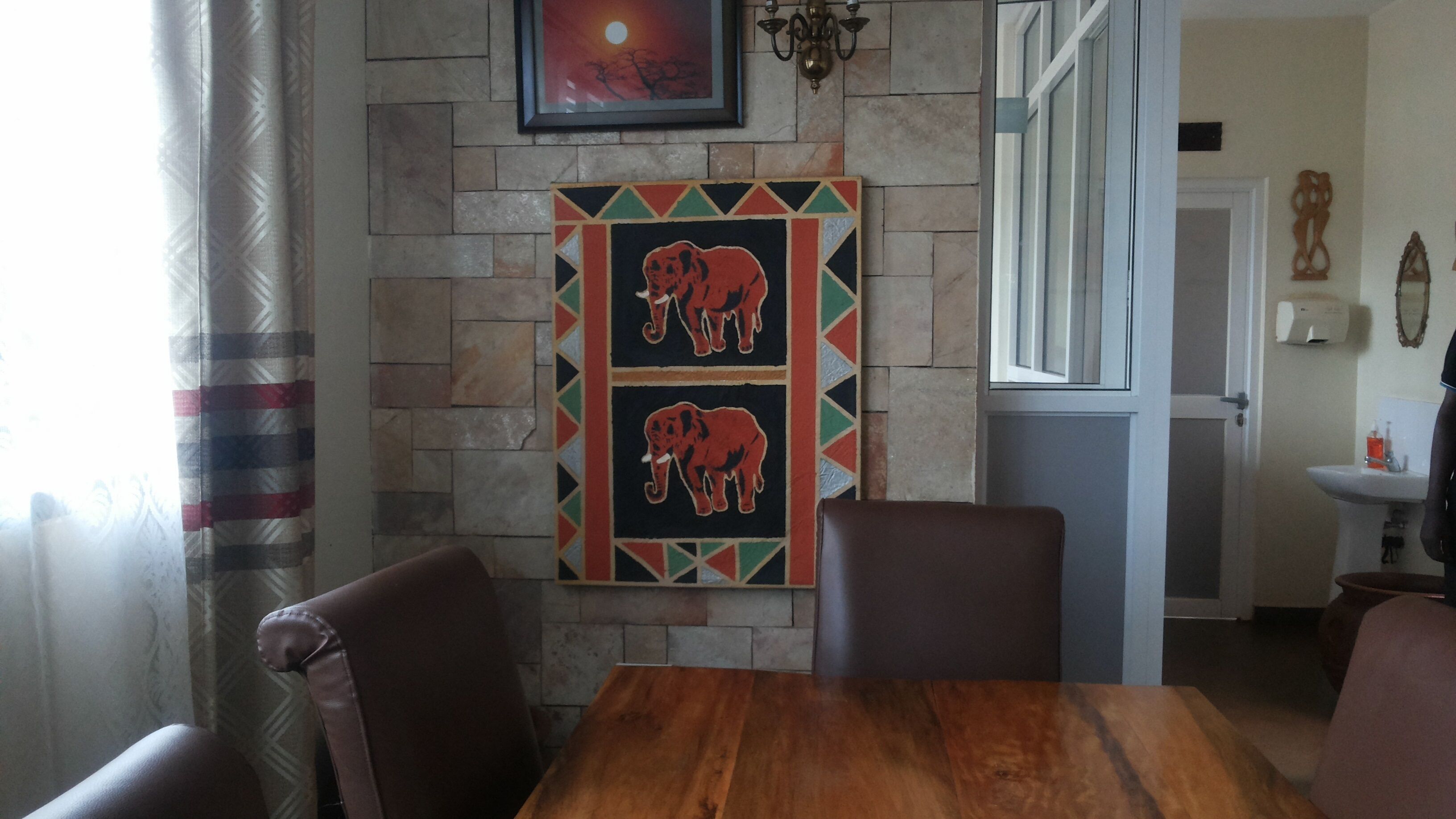 A dining area with a wooden table and chairs, a stone wall with a framed artwork of elephants, and a view into the adjacent room.