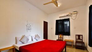 Quarto de hotel com uma cama, manta vermelha, ventoinha no teto, aparelho de ar condicionado, cadeira de madeira e uma pequena mesa.