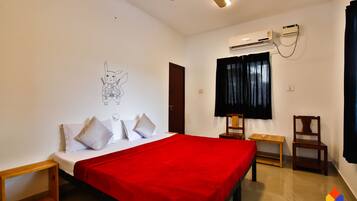 A hotel room with a bed, a red blanket, a ceiling fan, an air conditioning unit, a wooden chair, and a small table.