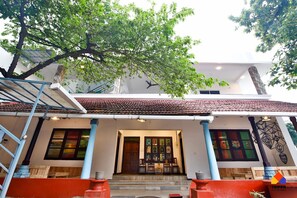 A two-story building with a tiled roof, a covered entrance with steps, and a large tree in front. - Trippr Alleppey - Backpacker Hostel (Ambalapuzha)