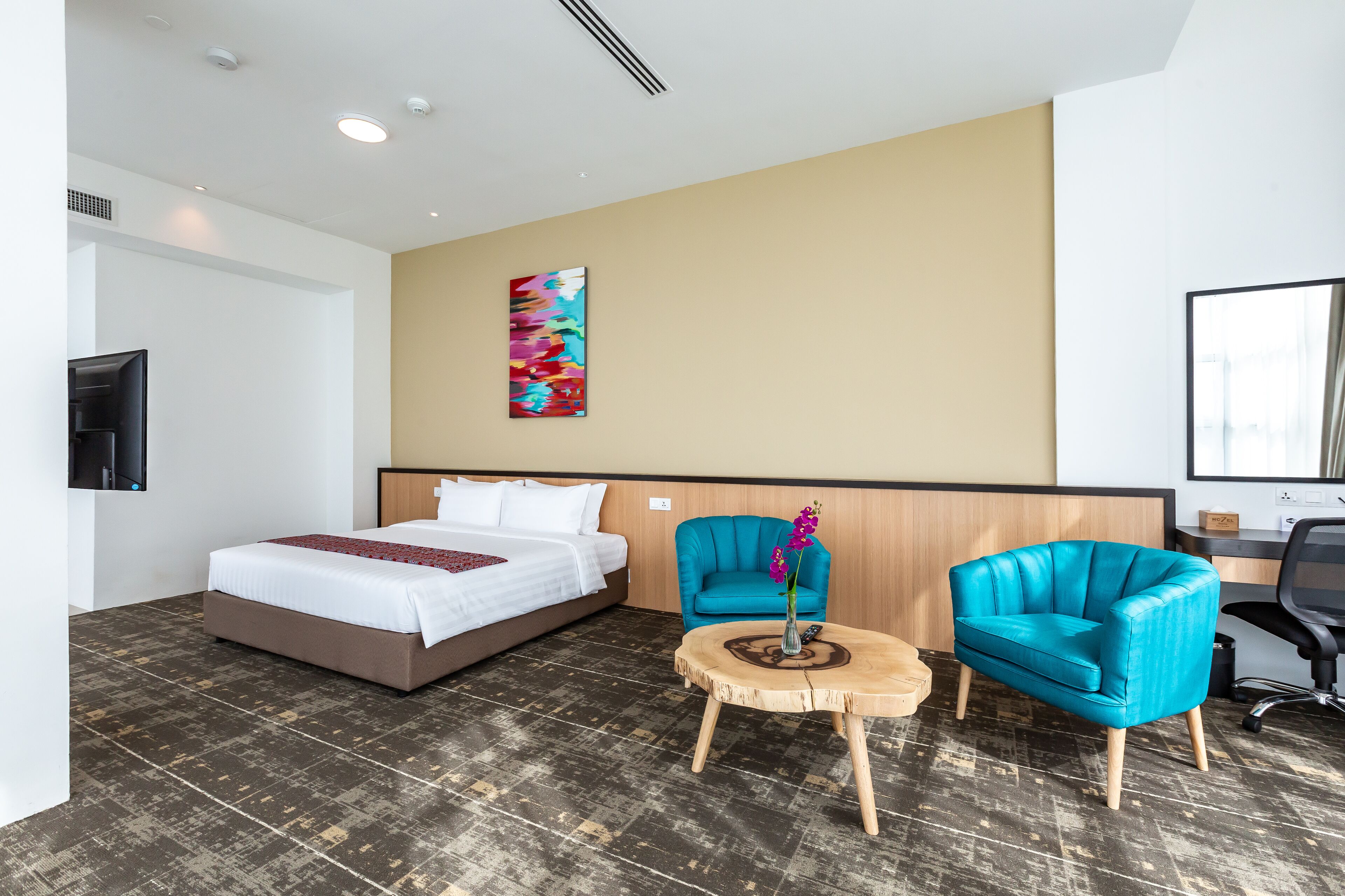 A hotel room with a bed, two chairs, a small table, a TV, and a colorful artwork on the wall.