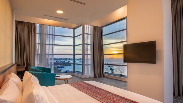 Een hotelkamer met een groot bed, een aan de muur bevestigde televisie, uitzicht op de zonsondergang over het water, en een balkon met een stoel.