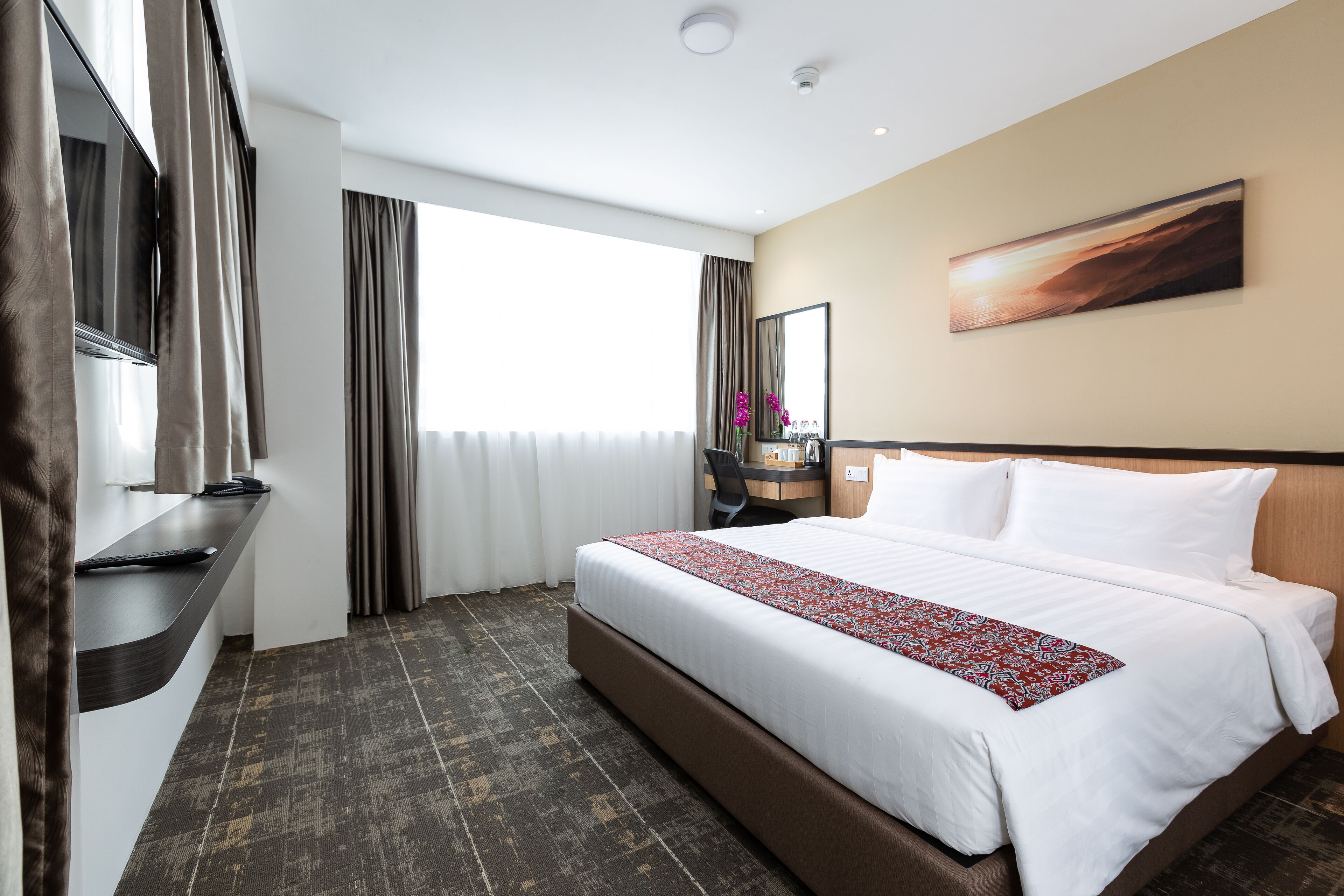 A hotel room with a large bed, a desk with a computer, a TV, and a window with curtains.