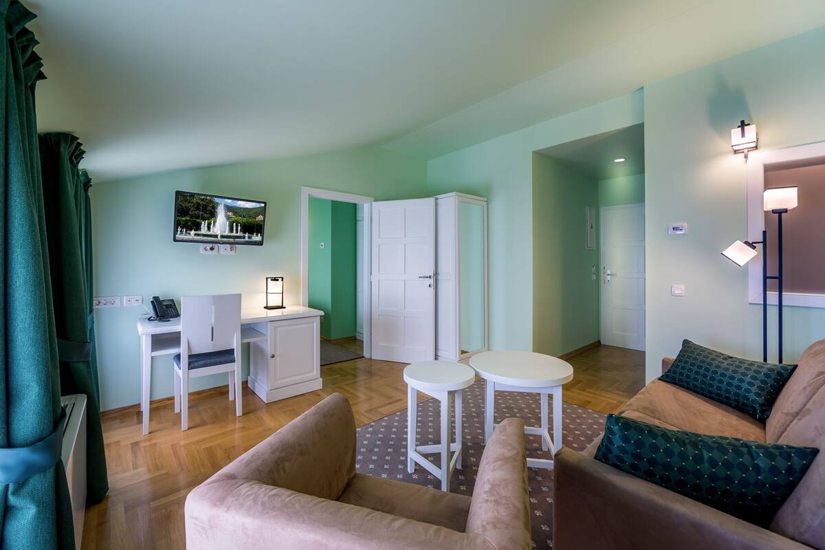 Ein modernes Wohnzimmer mit einer Couch, zwei kleinen Tischen, einem Schreibtisch mit Stuhl, einem an der Wand befestigten Fernseher und einem Kleiderschrank.