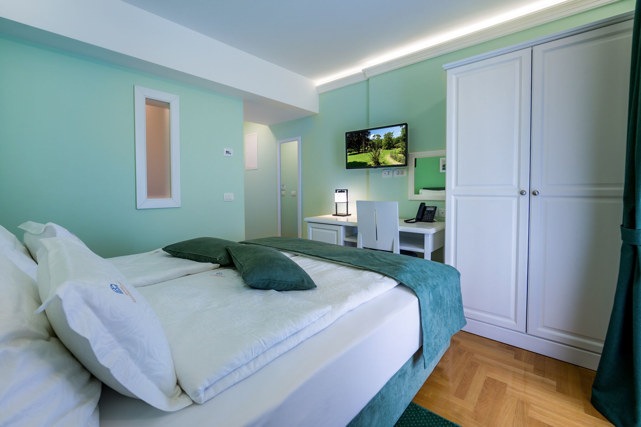 Un dormitorio ordenado con una cama, un televisor montado en la pared, un escritorio con una silla y un armario.