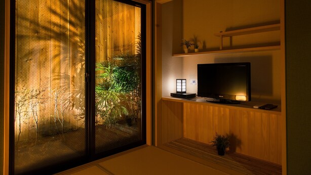 Et værelse med en skydedør til en have, et fladskærms-tv på en træhylde og et lille bord med en lampe.