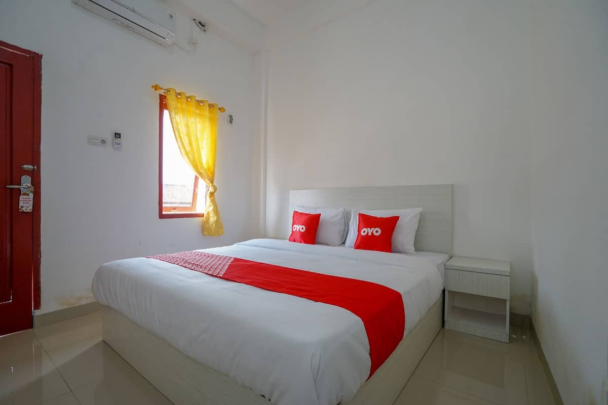 A hotel room with a large bed, white walls, a red door, and a window with yellow curtains.