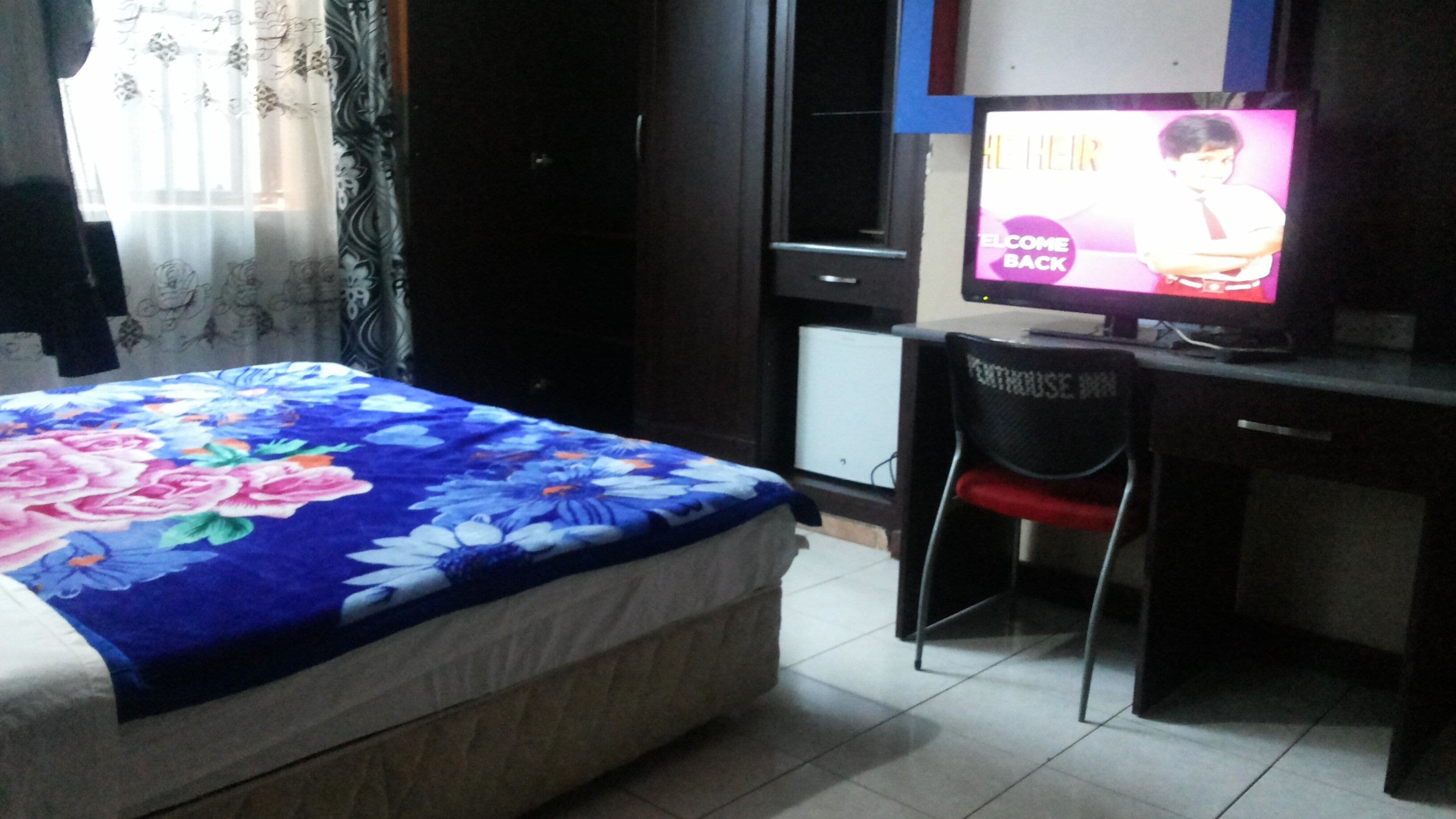 A bedroom with a bed, a desk, and a chair. There is a television on the desk displaying a welcome back message.
