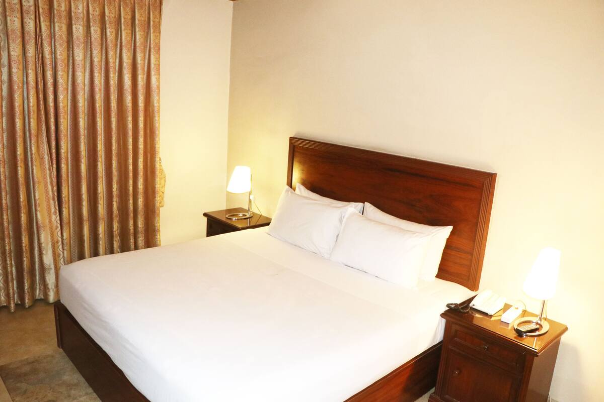 Habitación de hotel con una cama grande, dos mesitas de noche con lámparas, un cabecero de madera y una cortina.