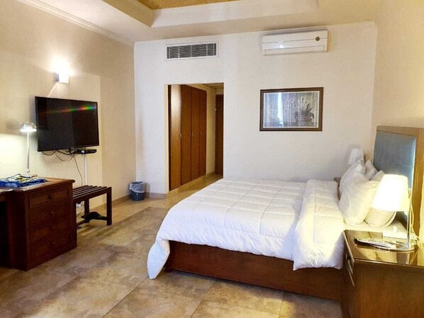 A bedroom with a bed, a desk, a television, and a door leading to another room.