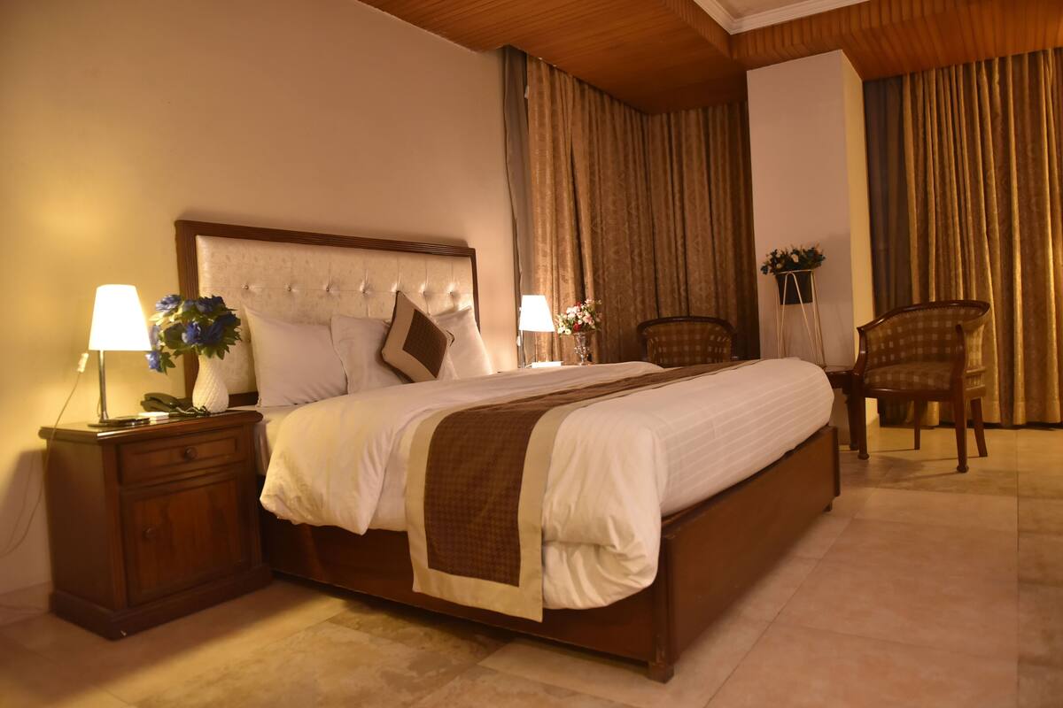 Una cama bien hecha con cabecera de madera, una mesita de noche con lámpara y una silla.
