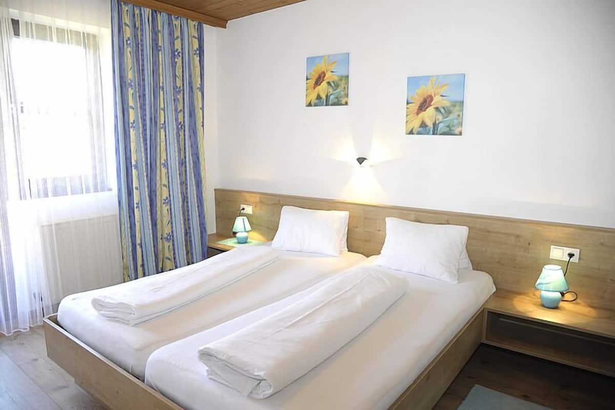 Una cama doble con ropa blanca, dos lámparas fijadas a la pared y dos girasoles enmarcados en la pared.