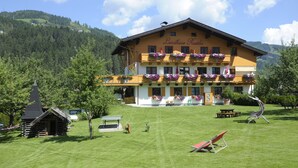A large two-story building with a sign reading "Landhaus Rüthka." It features balconies with flower boxes, a well-maintained lawn, and a small wooden structure in the foreground. - Landhaus & Haus Rustika (Wagrain)