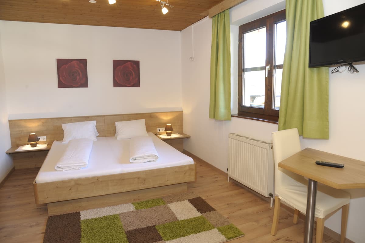 Un dormitorio con una cama de madera, dos mesitas de noche con lámparas, un radiador, un televisor montado en la pared y una ventana con cortinas verdes.