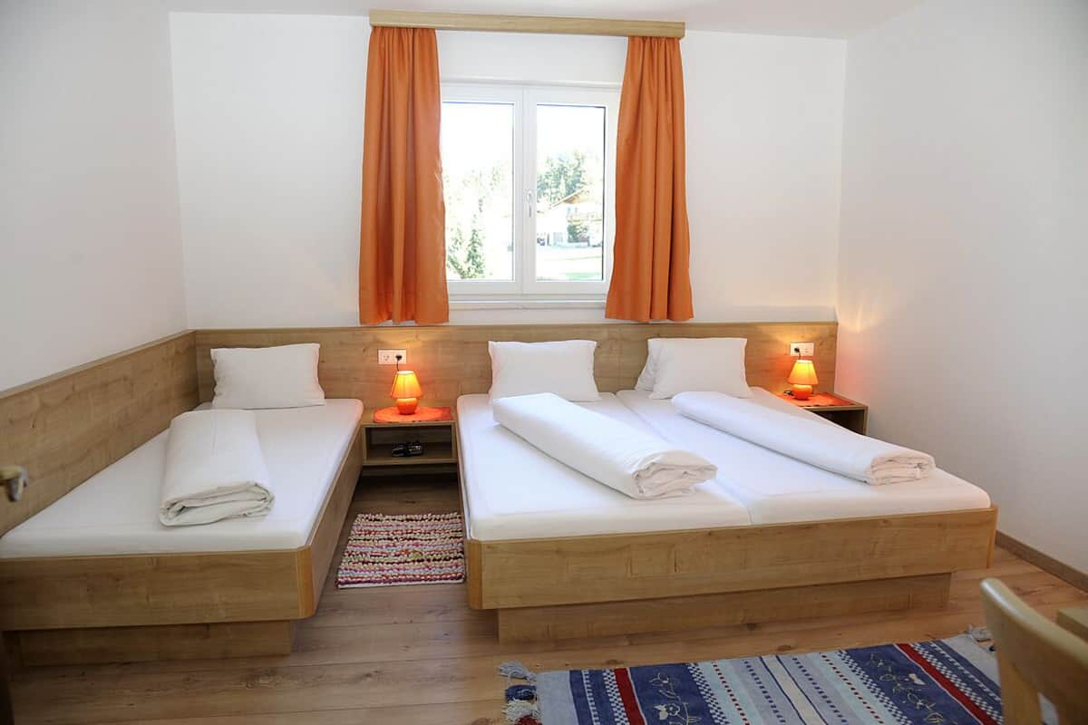 Una habitación con dos camas de madera, cada una con ropa blanca y almohadas, un ventanal con cortinas naranjas y una mesita de noche con una lámpara.