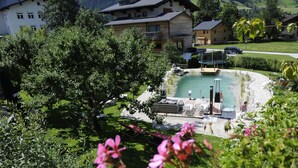 A pool area with lounge chairs and a small deck, surrounded by greenery and buildings in the background. - Landhaus & Haus Rustika (Wagrain)