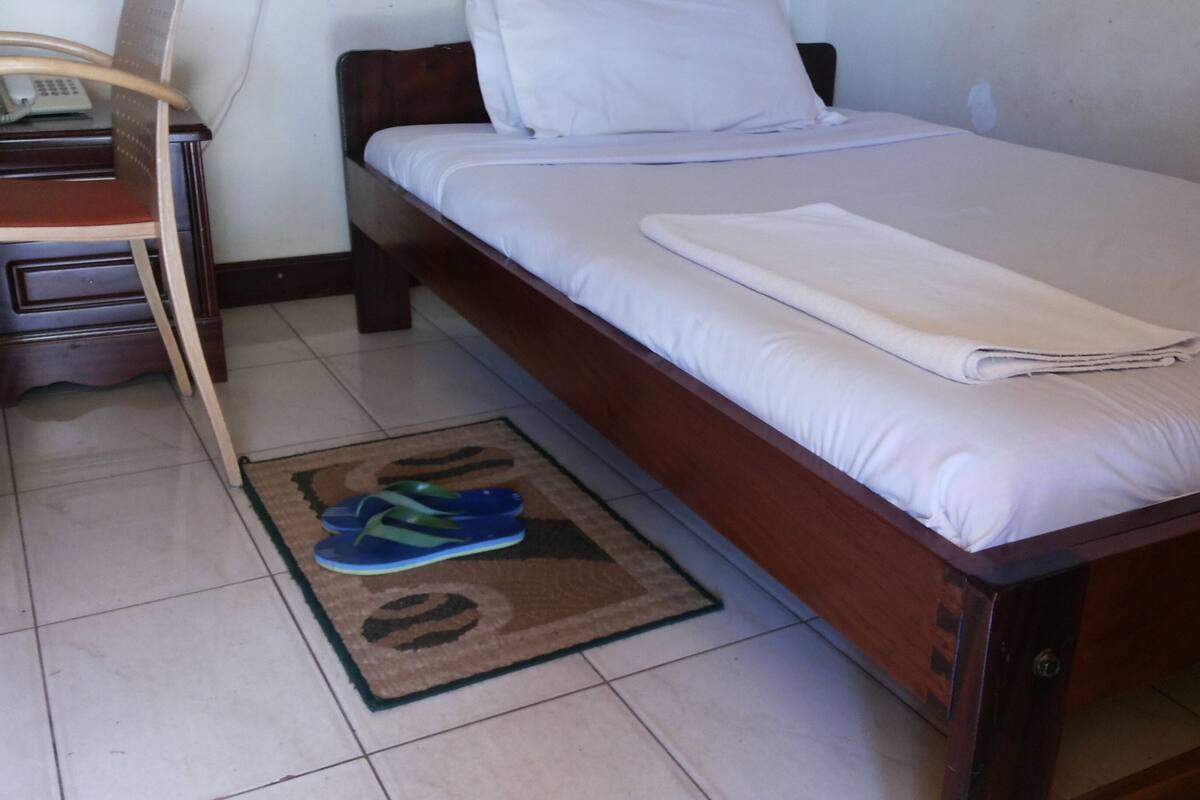 Ein Einzelbett mit weißer Matratze und Kissen, ein Holz-Kopfstück, ein kleiner Nachttisch mit Lampe sowie ein Paar blaue Flip-Flops auf einem Teppich.