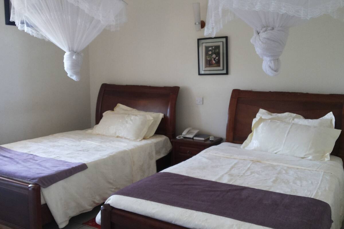 Ein Hotelzimmer mit zwei Betten, jeweils mit einem hölzernen Kopfteil und weißer Bettwäsche. An den Betten befinden sich Nachttische mit Telefonen und kleinen Tischen.