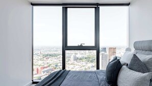 A bedroom with a large window, a bed with pillows, and a city view.