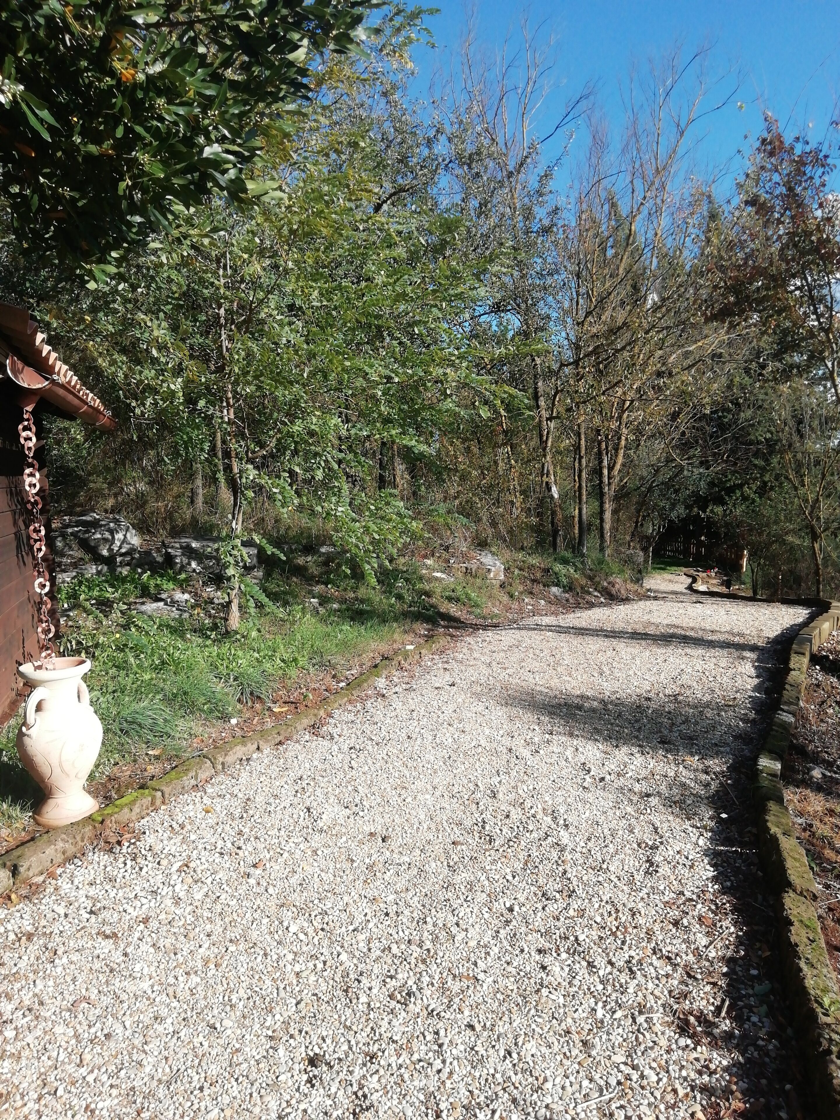 A gravel path leading through a wooded area with a rustic stone structure on the left.