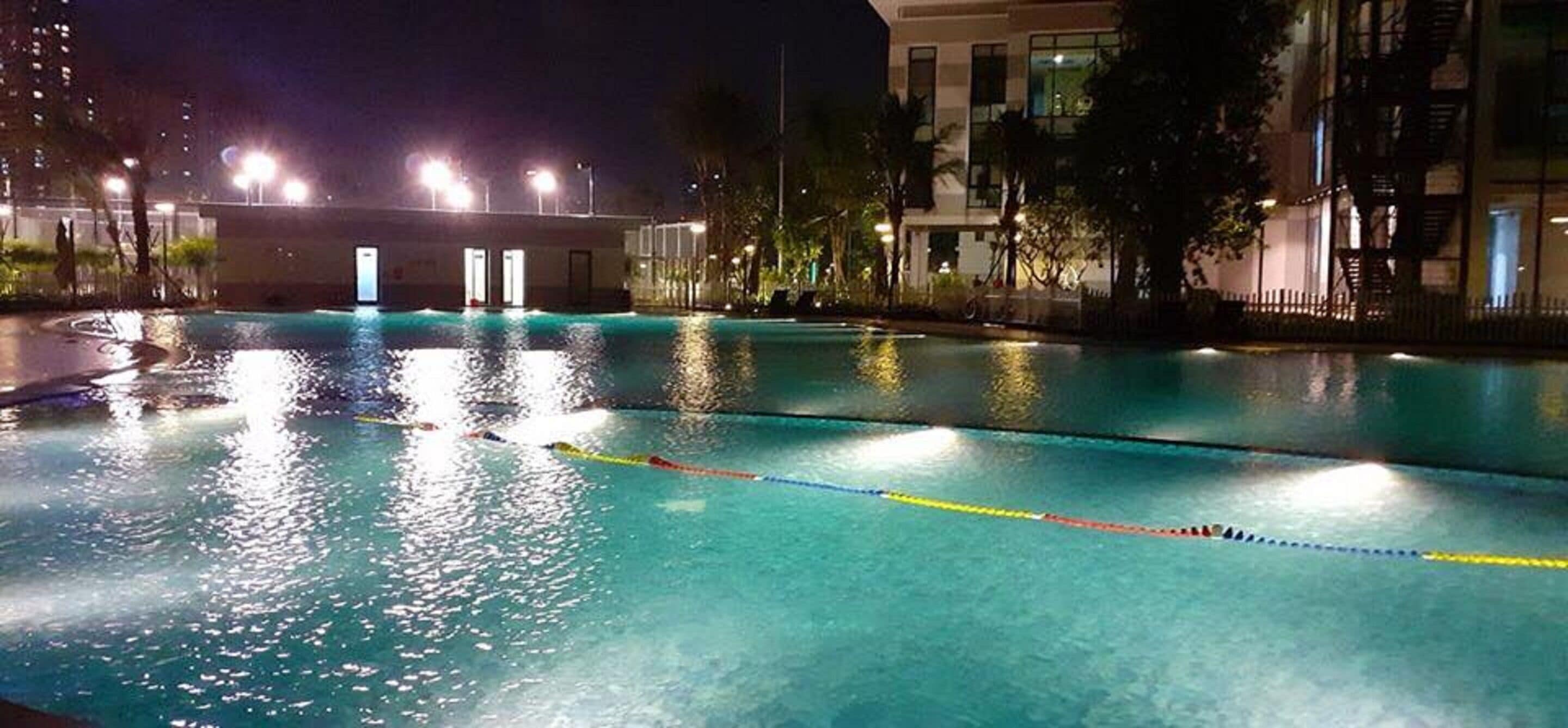 Une piscine la nuit, avec une eau claire, entourée de bâtiments et de palmiers.