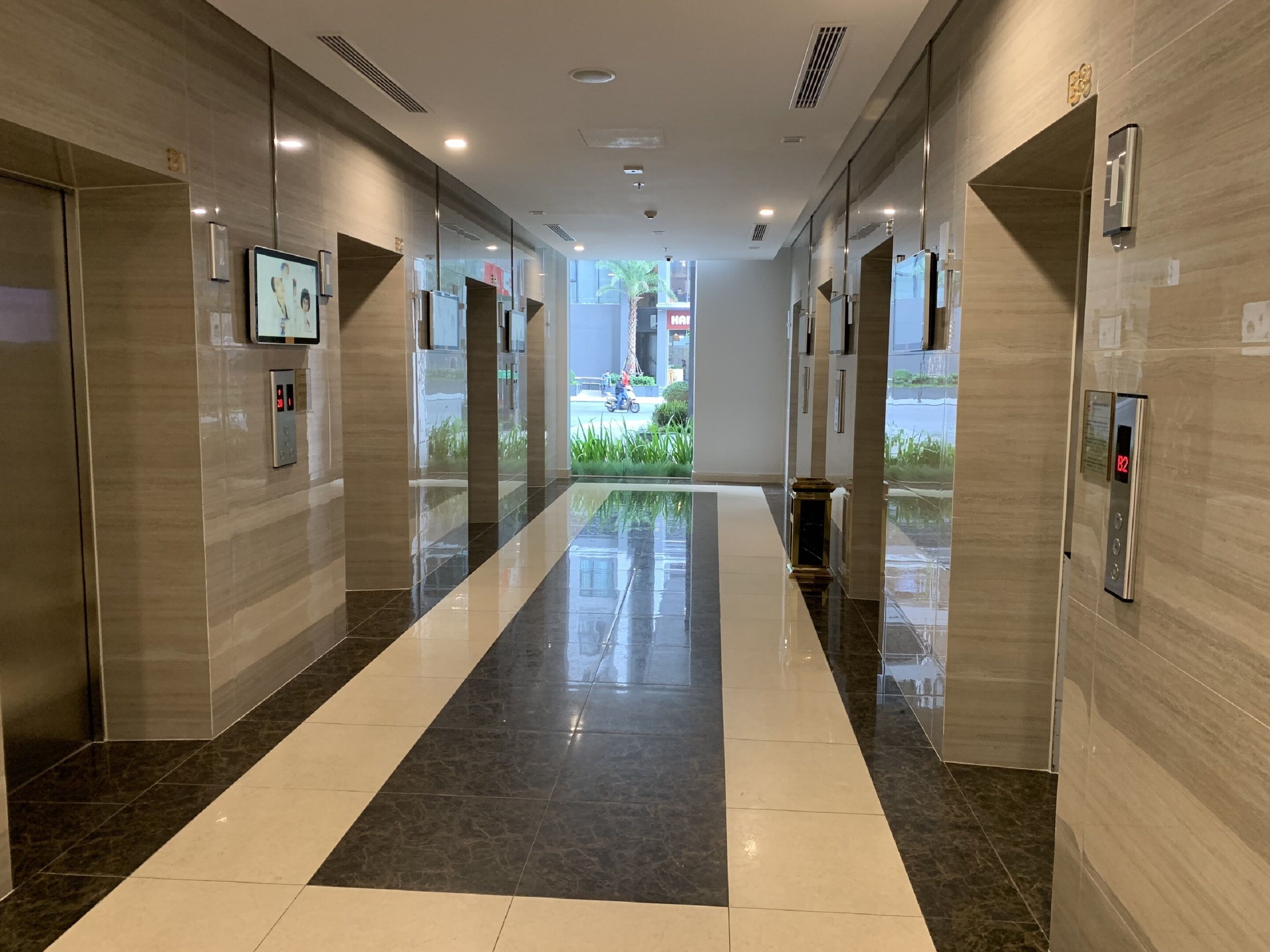 A modern hotel lobby with a reflective floor, elevator, and a large digital screen on the wall.