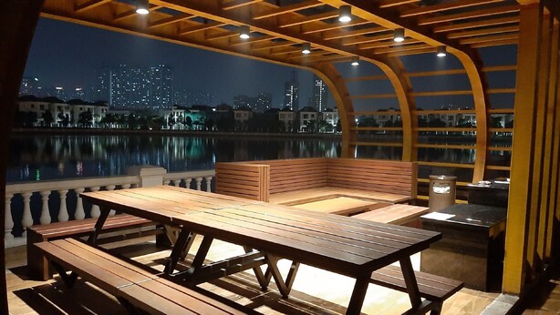 An outdoor seating area with wooden benches and tables, overlooking a body of water with a city skyline in the background. - Vinhomes Green Bay Apartments (Hanoi)