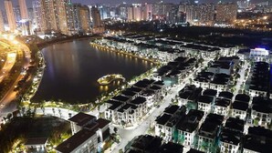A nighttime view of a cityscape with high-rise buildings, a body of water, and a residential area with streets and buildings. - Vinhomes Green Bay Apartments (Hanoi)