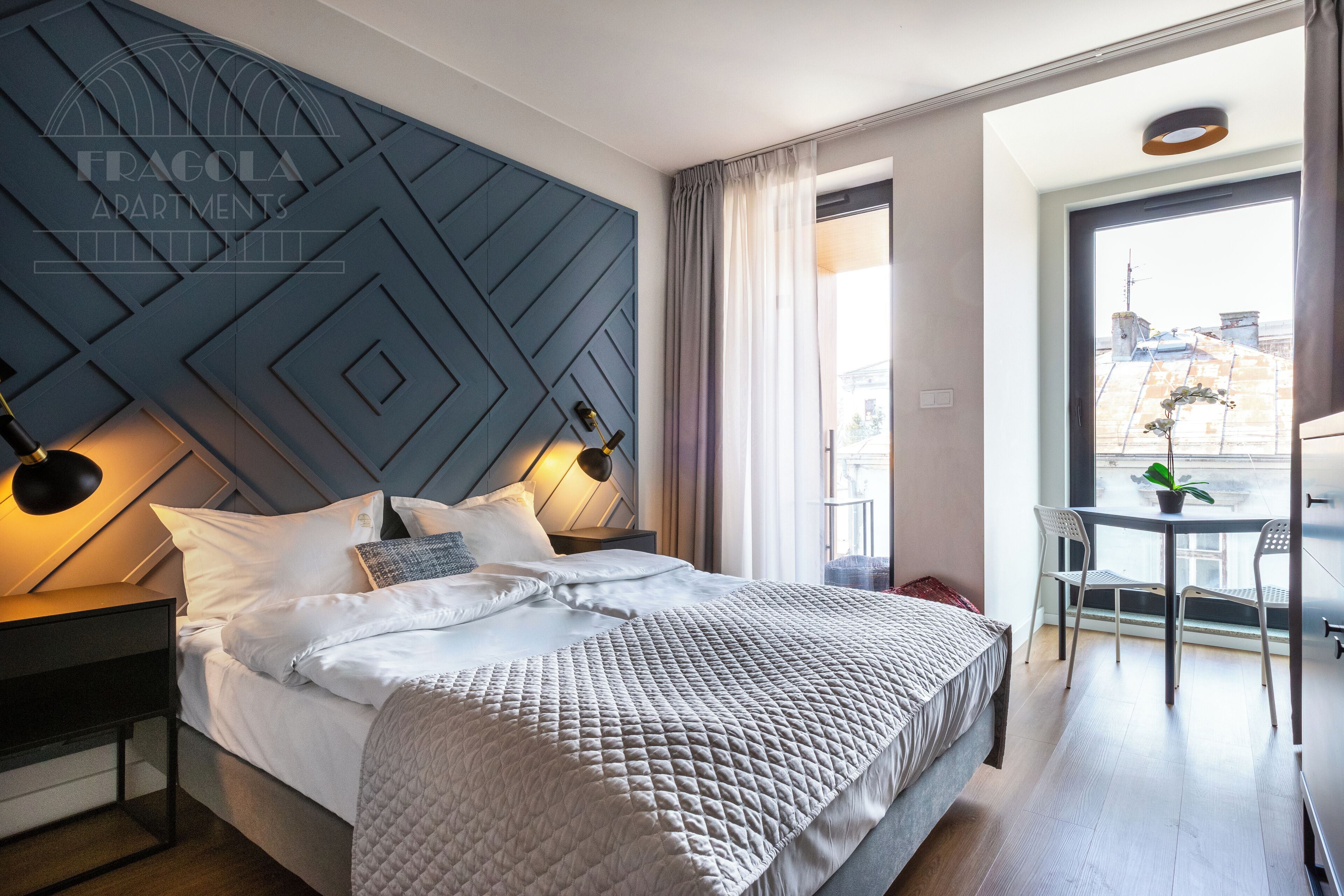 Un dormitorio moderno con una cama grande, un cabecero con diseños geométricos, lámparas de noche, una pequeña mesa con sillas y vistas a la ciudad a través de un ventanal amplio.