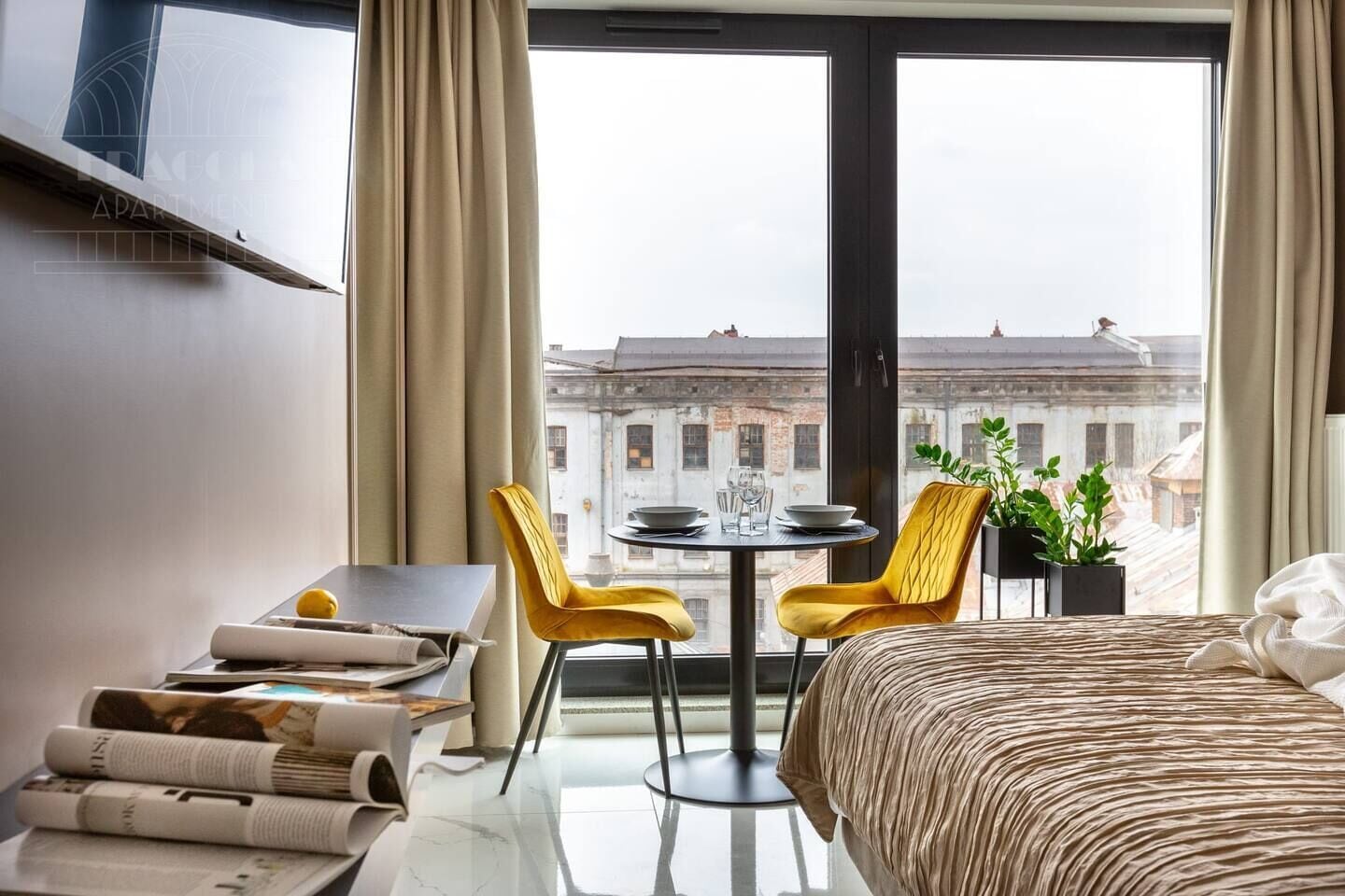 A hotel room with a bed, a dining table with chairs, a small balcony, and a view of buildings.